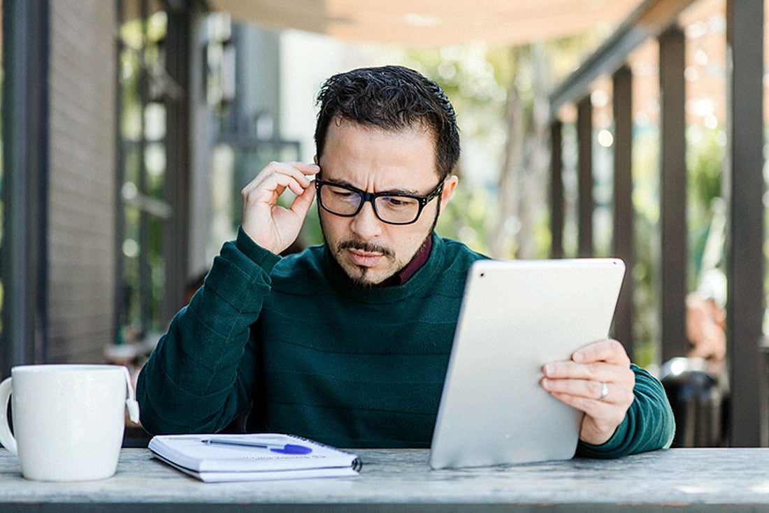 Middle-aged man with glasses holding tablet and squinting at text, indicating presbyopia vision difficulty or dry eye.