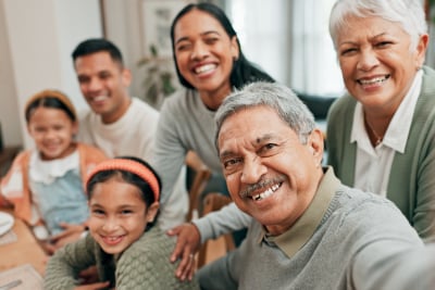 Smiling multi-generational family gathered around a table, with a grandfather taking a close-up selfie.