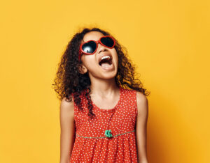 Young girl in red sunglasses and dress smiling, representing UV protection for children’s eye health