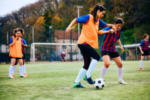 Girls playing soccer on a green field, one in an orange bib dribbling the ball as teammates and opponents watch near a goal.