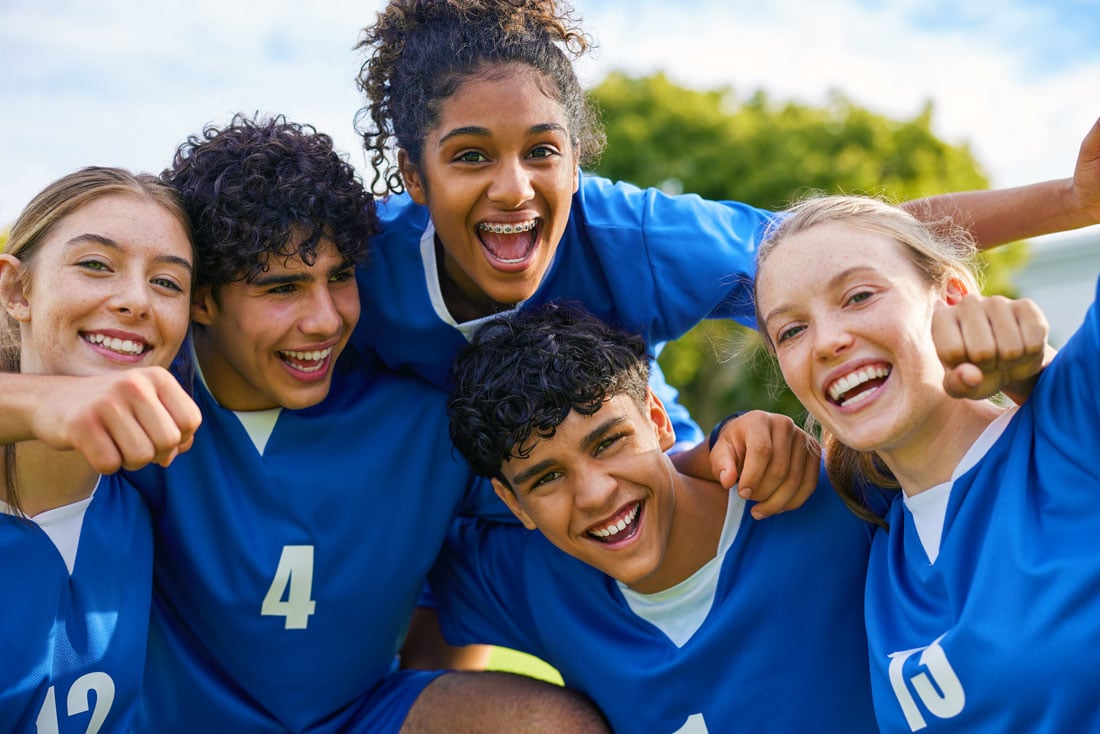 A diverse group of smiling young athletes in blue jerseys huddle together outdoors.