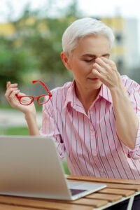 An older woman with short white hair wearing a pink striped shirt sits at a wooden table with a laptop, holding red glasses and pinching her nose.
