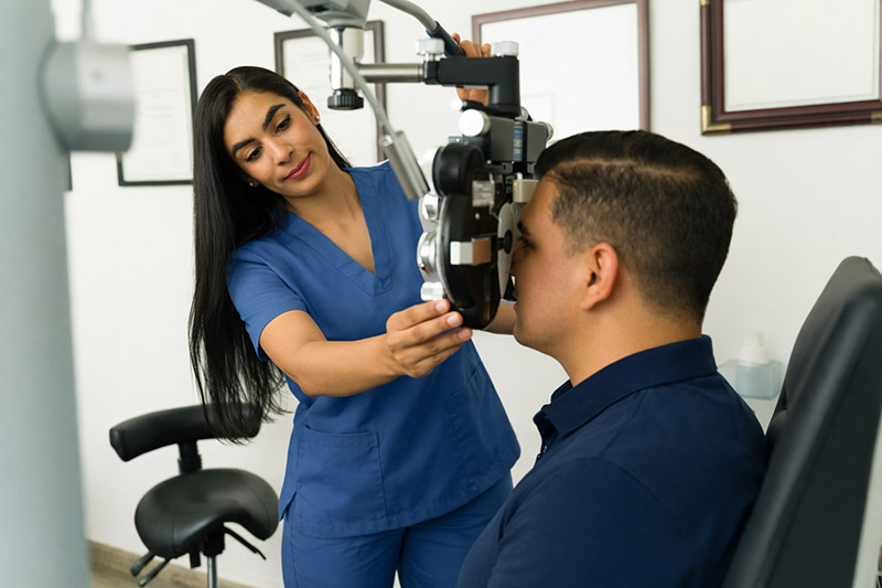 Prepare for your appointment with a friendly eye care professional adjusting a phoropter during an in-office eye exam, as a patient in a navy collared polo sits comfortably in the exam chair.