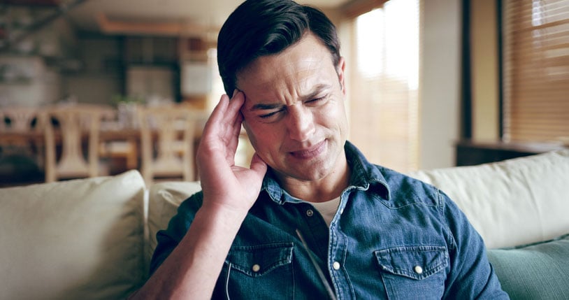 Man sitting indoors with eyes closed and hand to his temple, appearing sensitive to light or experiencing visual disturbance associated with an ocular migraine