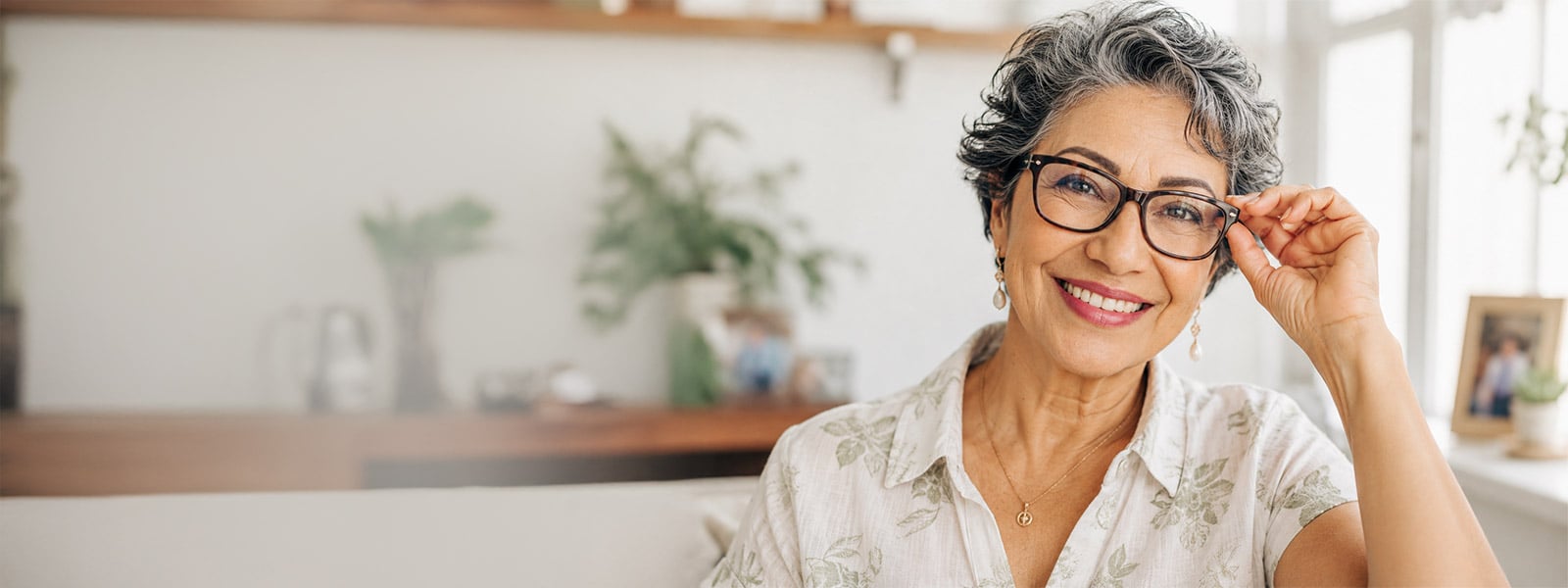 Smiling older Mexican woman wearing glasses and a light summer shirt, representing friendly eye care in a bright home setting.